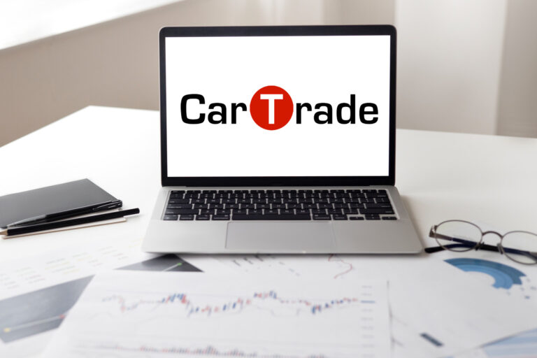 Image representing a pen, glasses and papers representing CarTrade Logo on Laptop
