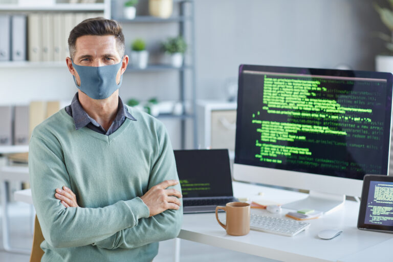 Portrait of mature businessman in protective mask standing with arms crossed and looking at camera working at IT office
