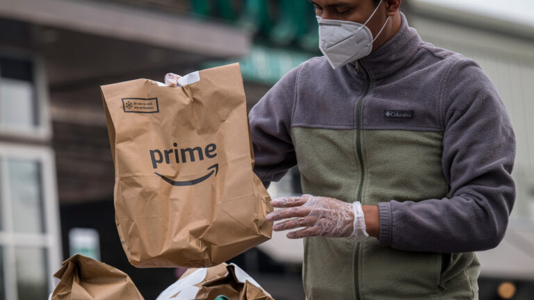 An independent contractor wearing a protective mask and gloves loads Amazon Prime grocery bags into a car outside a Whole Foods Market