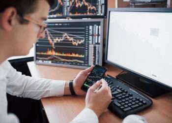 Over the shoulder view of and stock broker trading online while accepting orders by phone. Multiple computer screens ful of charts and data analyses in background.