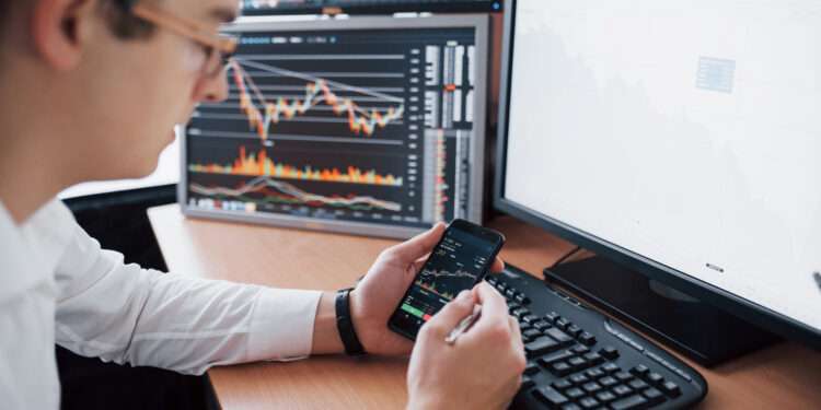 Over the shoulder view of and stock broker trading online while accepting orders by phone. Multiple computer screens ful of charts and data analyses in background.